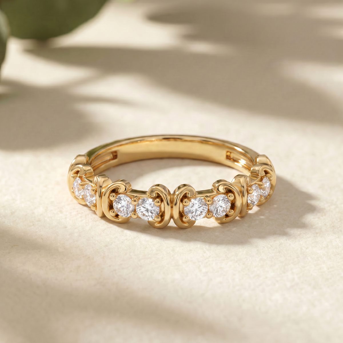 A gold eternity-style ring with small clear gemstones set in an ornate repeating pattern, photographed on a warm beige surface with soft leaf shadows.
