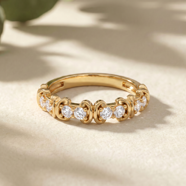 A gold eternity-style ring with small clear gemstones set in an ornate repeating pattern, photographed on a warm beige surface with soft leaf shadows.