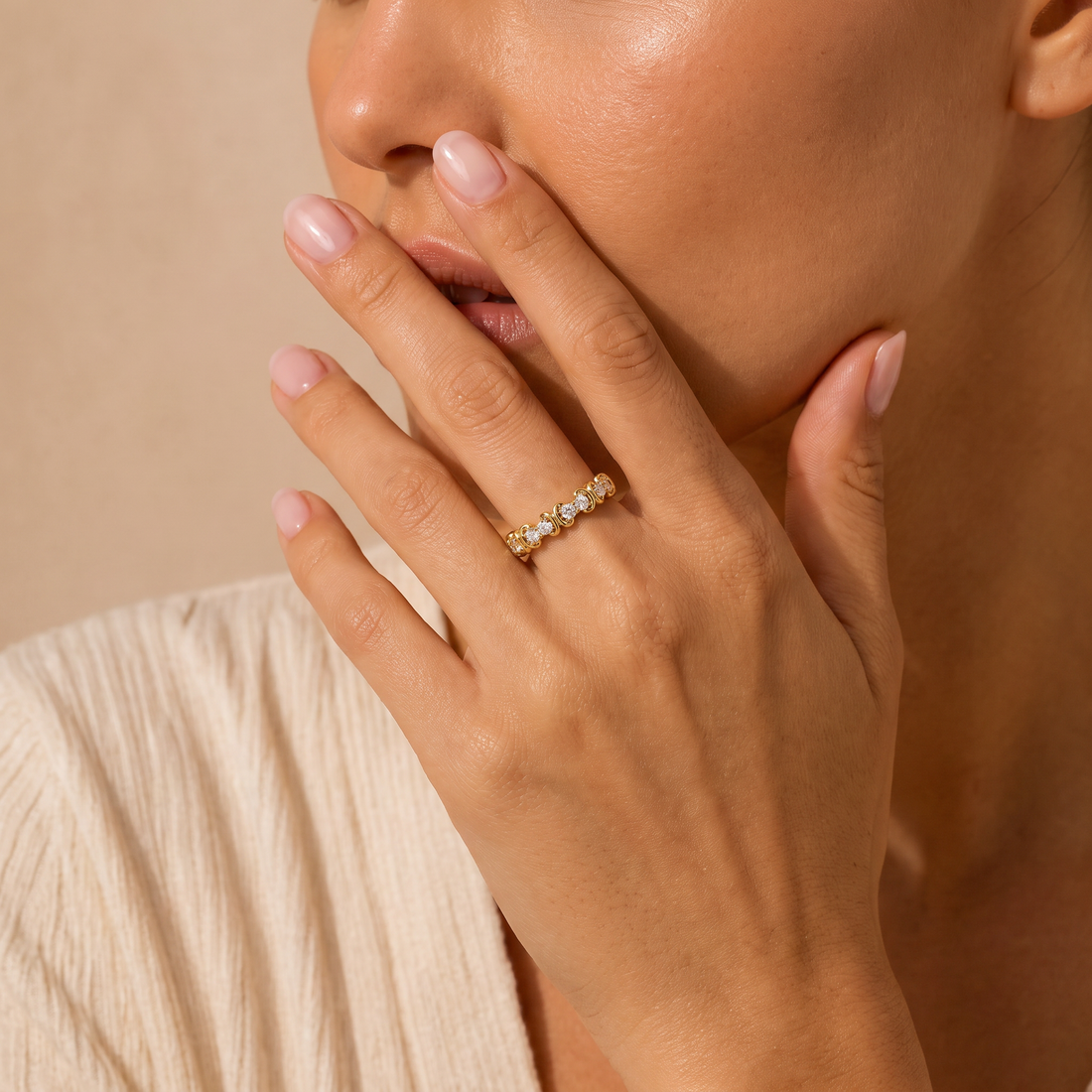 A close-up lifestyle shot of a woman wearing a delicate gold band ring set with small clear stones, resting on her ring finger. She is wearing a cream-colored textured top against a warm beige background.