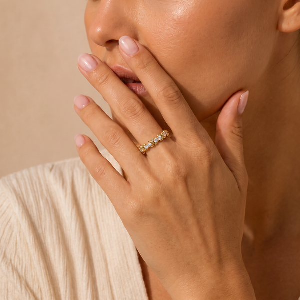 A close-up lifestyle shot of a woman wearing a delicate gold band ring set with small clear stones, resting on her ring finger. She is wearing a cream-colored textured top against a warm beige background.