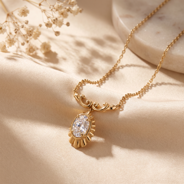 A gold chain necklace with an ornate oval pendant featuring a clear faceted gemstone, styled on draped beige fabric with dried flowers and a marble slab.