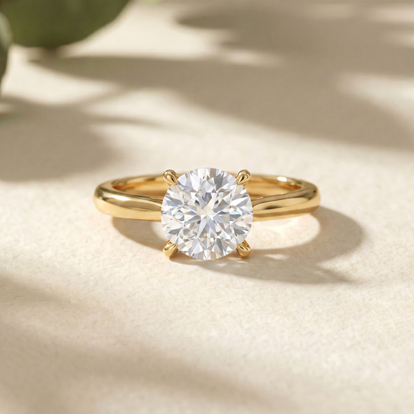 A round-cut diamond solitaire ring set in a polished yellow gold band, resting on a textured beige surface with soft leaf shadows.
