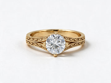 A yellow gold solitaire engagement ring with an intricate filigree band and a round brilliant-cut diamond center stone, shown on a clean white background.