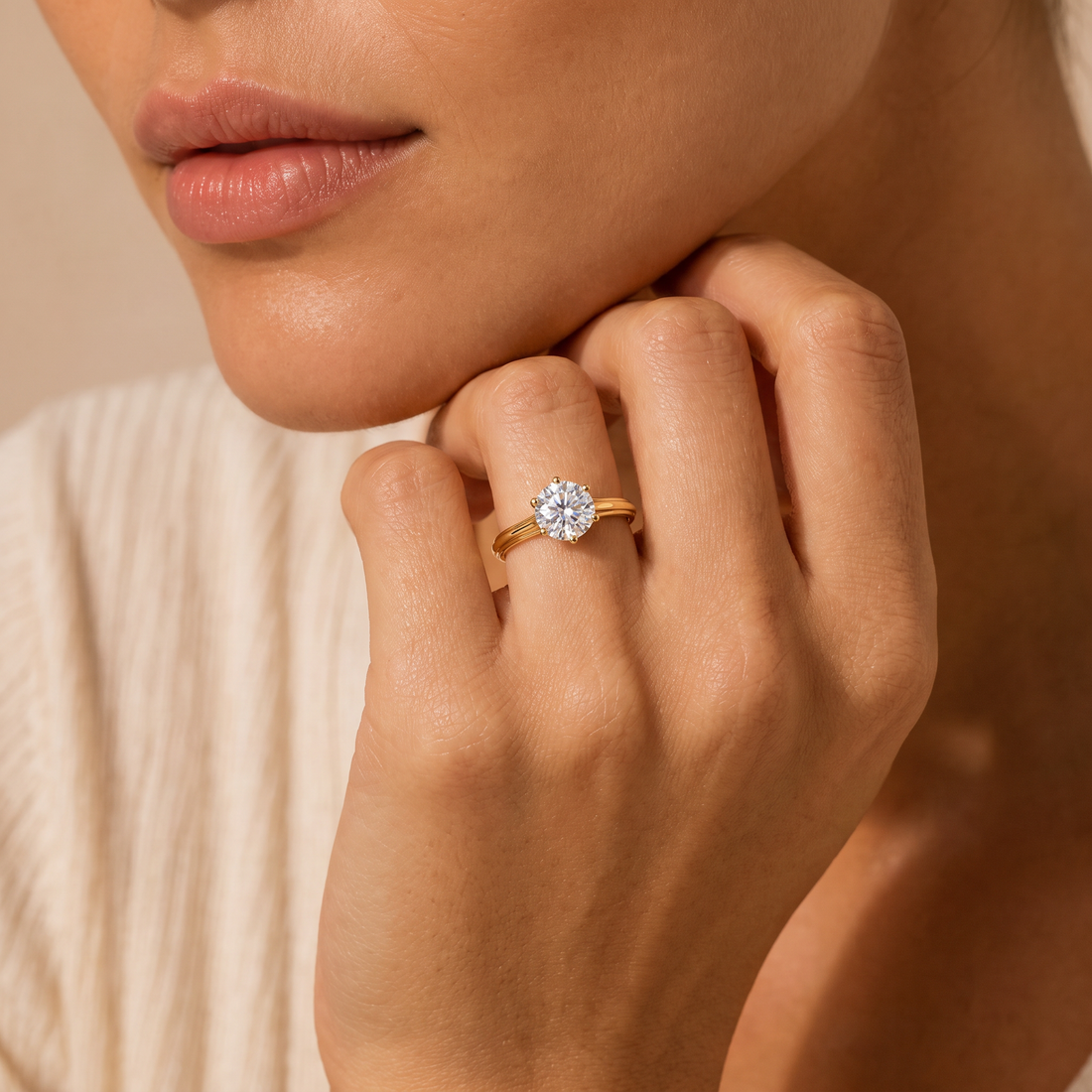A close-up lifestyle shot of a woman wearing a yellow gold engagement ring with a round brilliant-cut diamond on her ring finger. The background is a soft, warm beige tone.