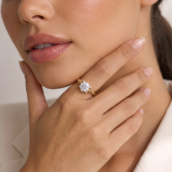 A close-up lifestyle shot of a model wearing a yellow gold ring with a round-cut clear gemstone on her finger, set against a soft, light beige background.