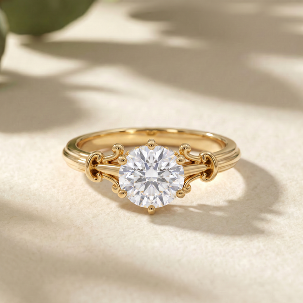 A gold solitaire engagement ring with a round brilliant-cut clear diamonds, set in a delicate three-prong setting with decorative scrollwork on the band, resting on a textured beige surface with soft leaf shadows.