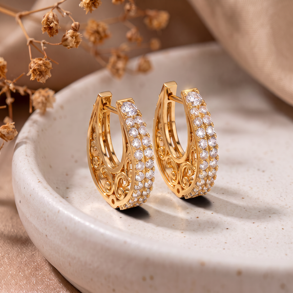 A pair of gold huggie hoop earrings adorned with rows of clear pavé stones and intricate filigree detailing, displayed on a light beige ceramic dish with dried flowers in the background.