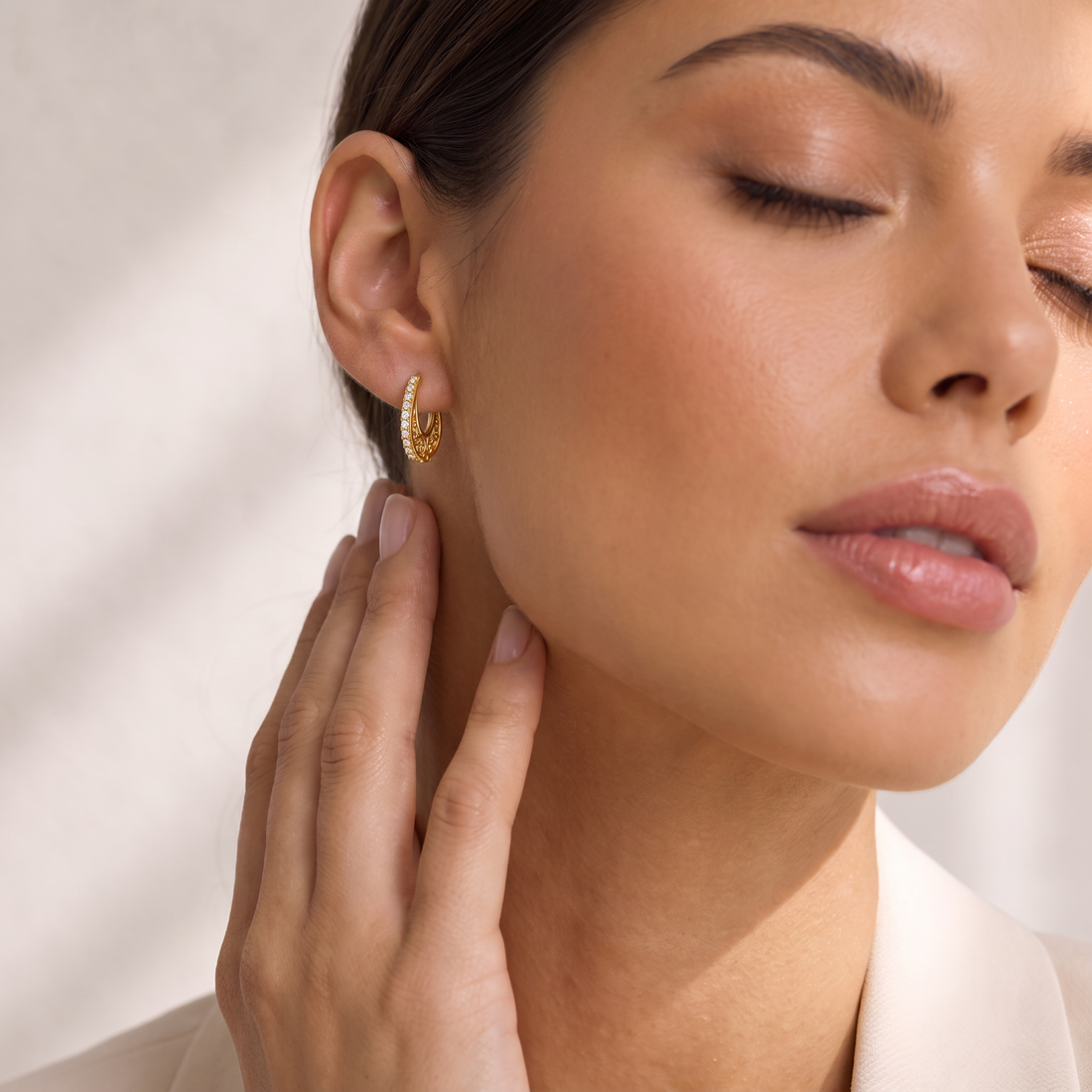 A close-up lifestyle shot of a woman wearing a small gold crescent-shaped hoop earring with a textured, beaded outer edge. She is shown from the side with her hand resting on her neck, wearing a light-colored collared shirt against a softly lit, neutral background.